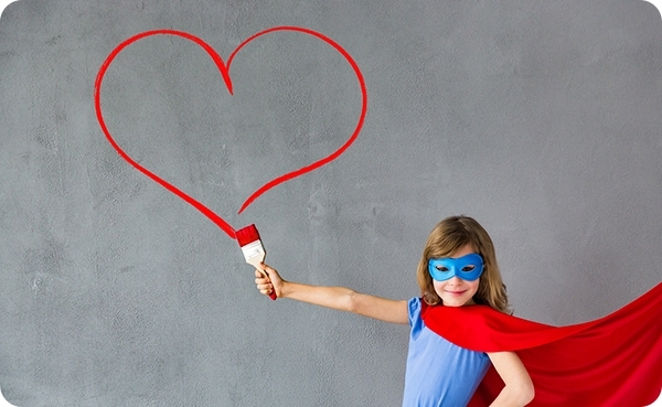 A girl dressed up in a red cape and a blue mask holds a paintbrush. On the wall marks a red heart that the girl painted.